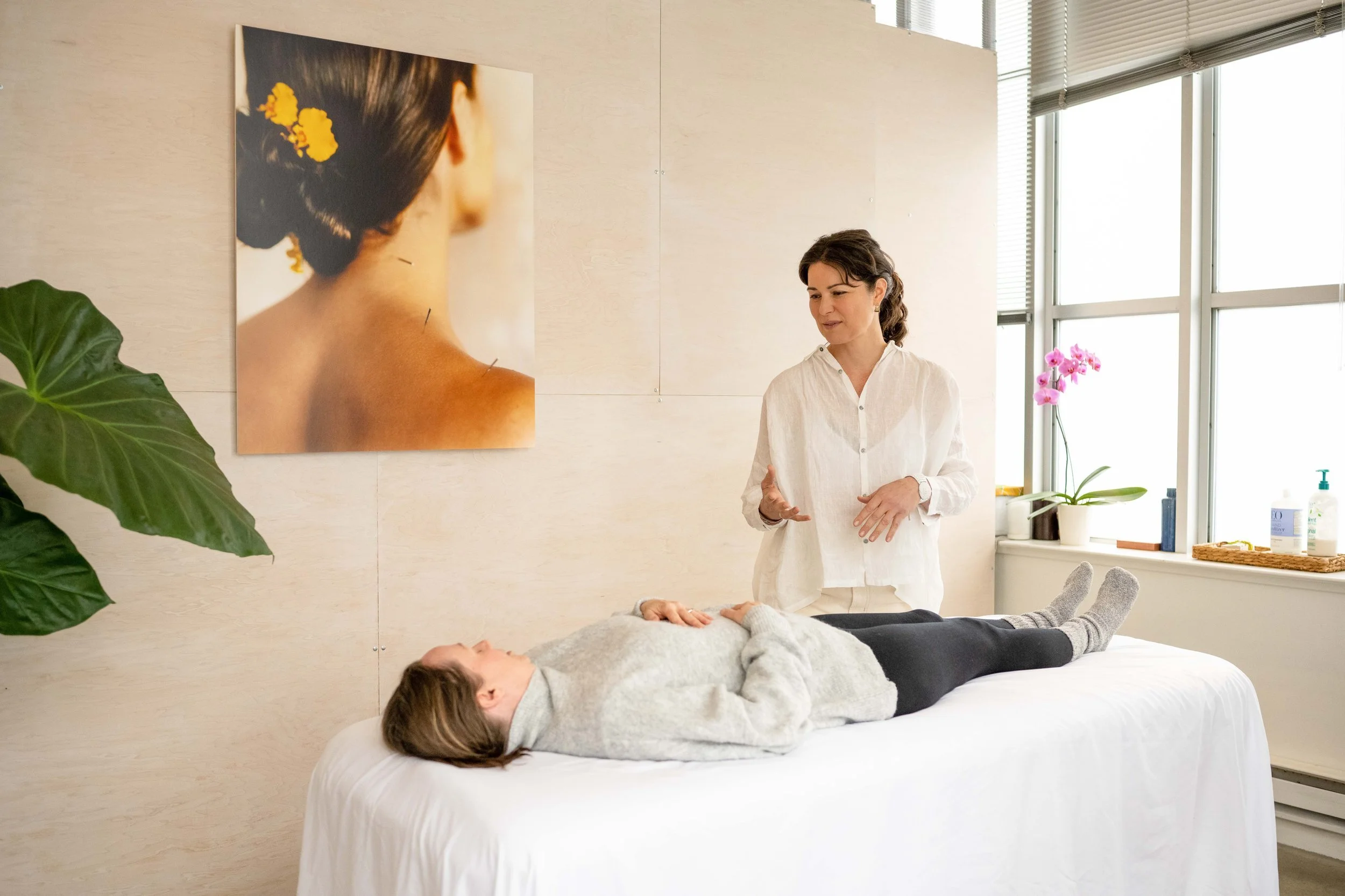 A woman giving a massage to a person lying on a bed in a bright room with large windows, plants, and artwork on the wall.