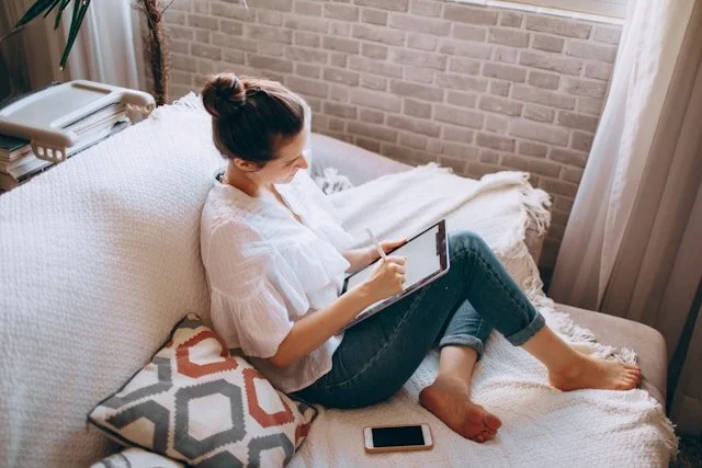 Woman sitting on a bed using a tablet, with a smartphone on the bed beside her, in a cozy bedroom with a textured wall and a window with curtains.