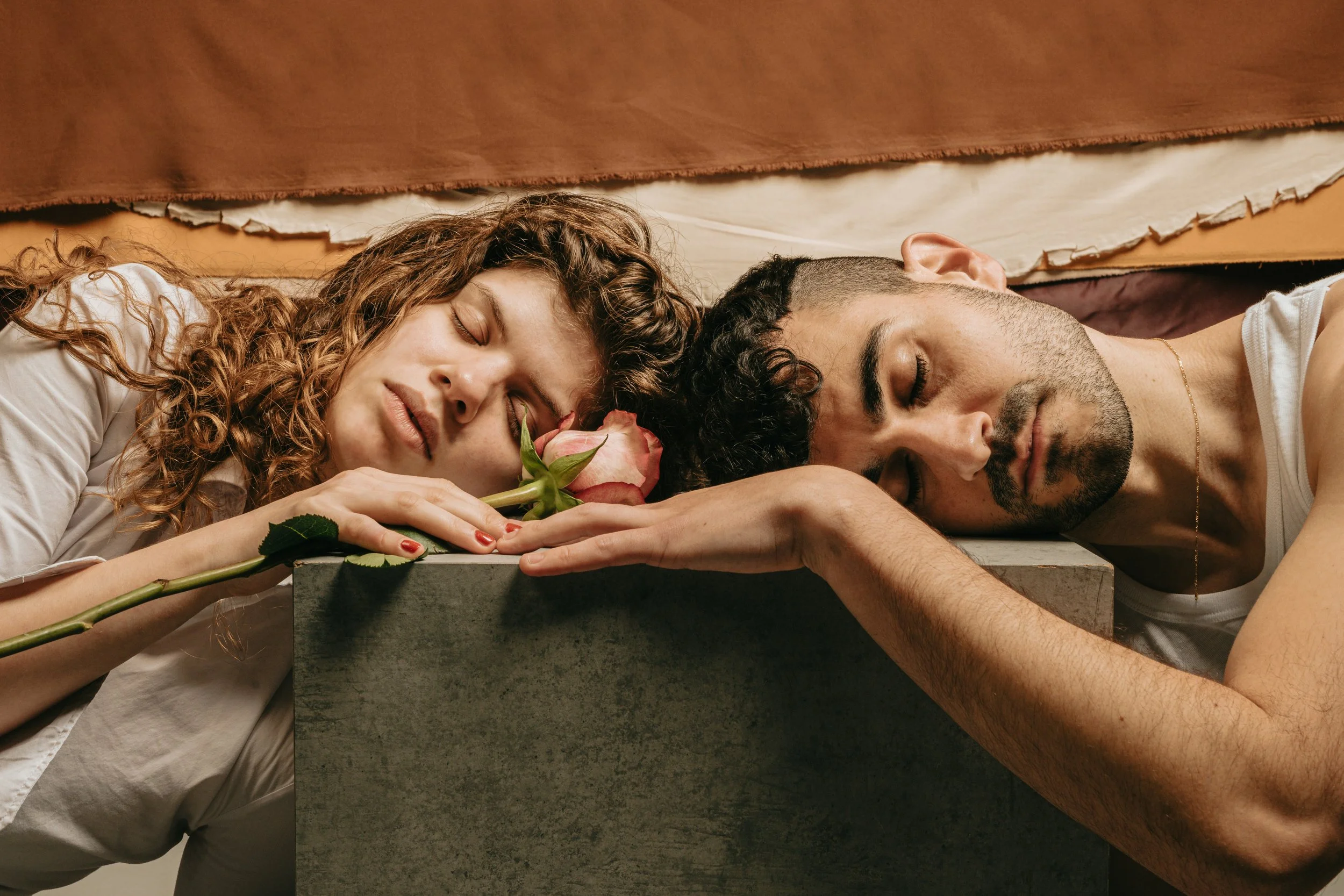 A woman with curly hair and a man with dark curly hair and a beard are lying on a surface, with their heads touching a concrete block. The woman is holding a pink rose near her face while resting her cheek against it. Both have their eyes closed and appear to be peacefully sleeping.