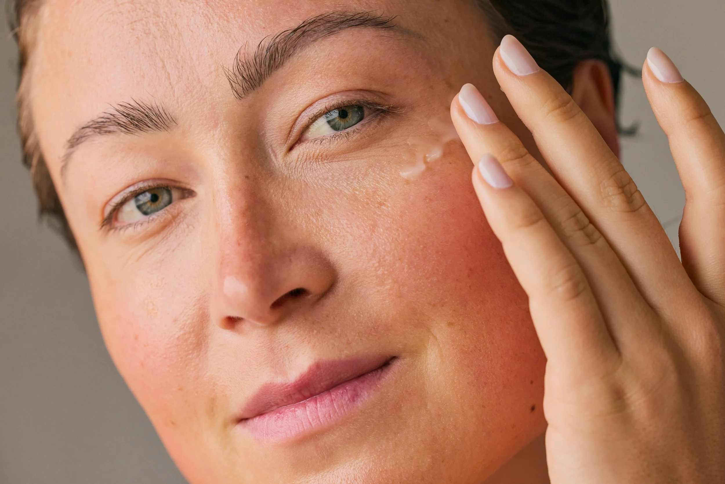 Close-up of a woman's face with her hand touching her temple, showing clear skin, blue eyes, and well-groomed eyebrows.