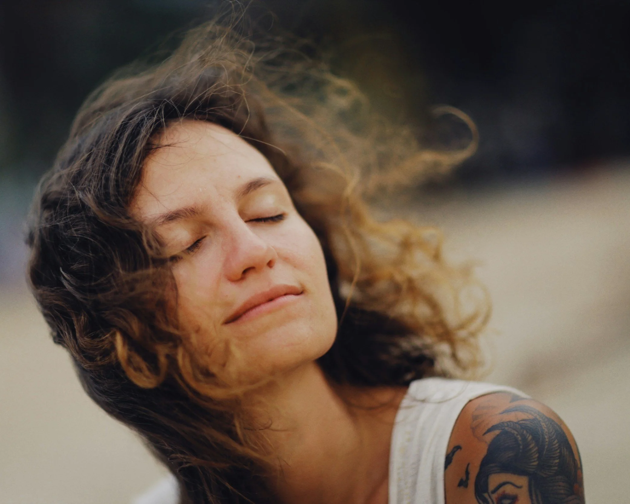 Close-up of a woman with closed eyes, curly hair blowing in the wind, and a tattoo on her shoulder, against a neutral background.