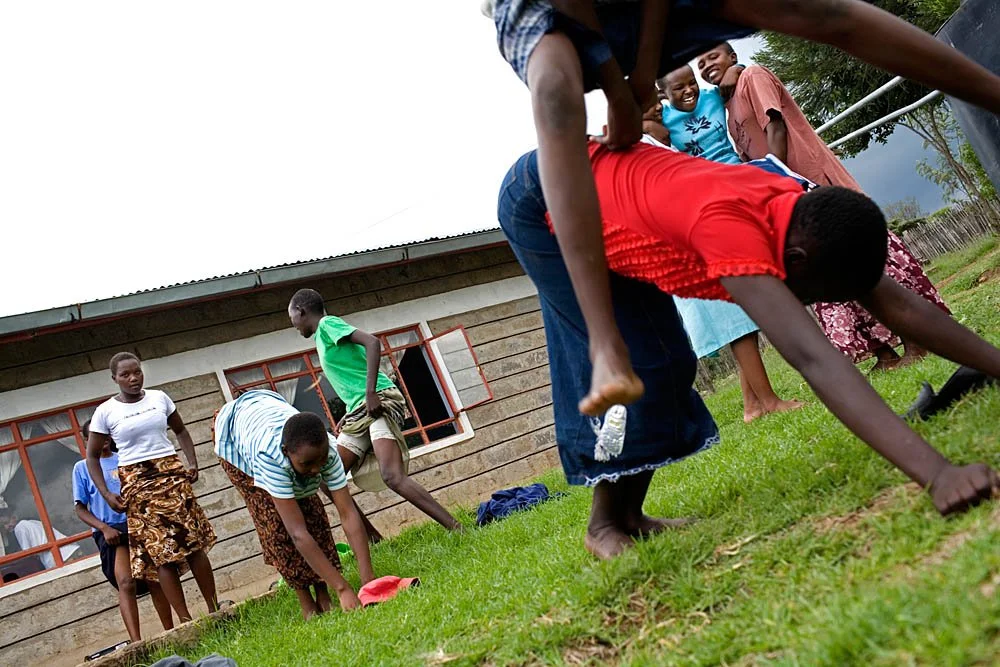 Group of children and adults playing on grass outside a building, with some participants crawling and others observing, in a rural setting.