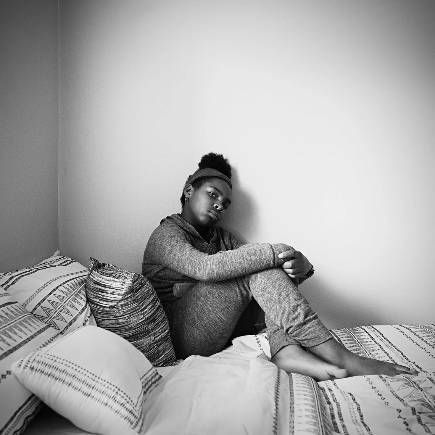Black and white photo of a woman with her hair in a bun, sitting on a bed with patterned pillows, looking at the camera with a serious expression.