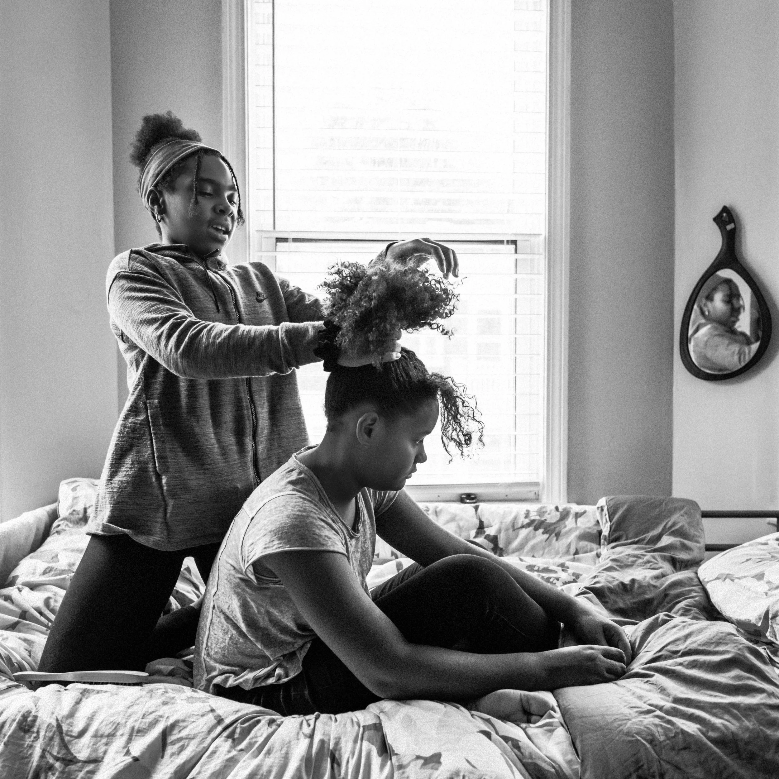 Two women styling curly hair on a bed in a bedroom, with a window and mirror visible in the background.