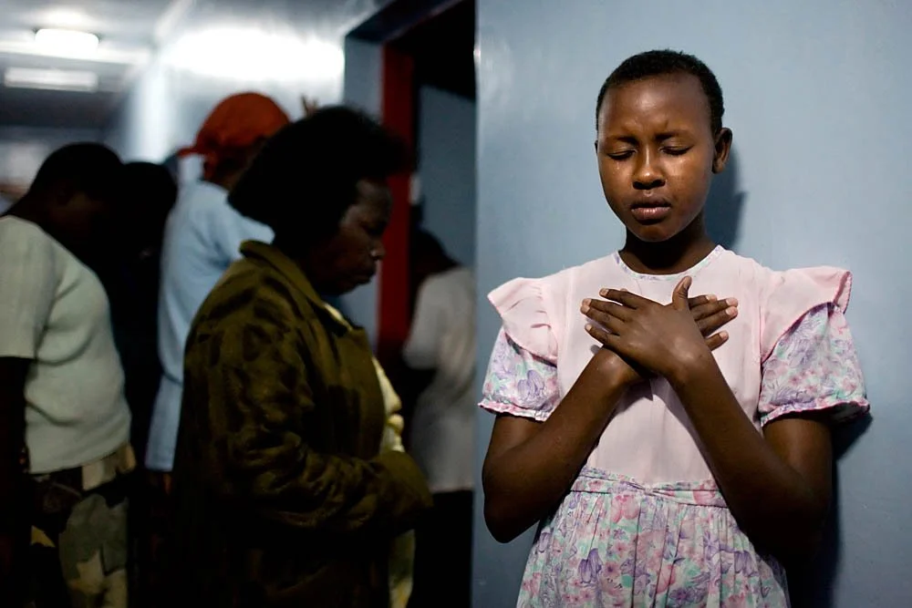 A young girl with closed eyes and hands on her chest, appearing to pray or reflect, in a room with a group of people standing in the background.