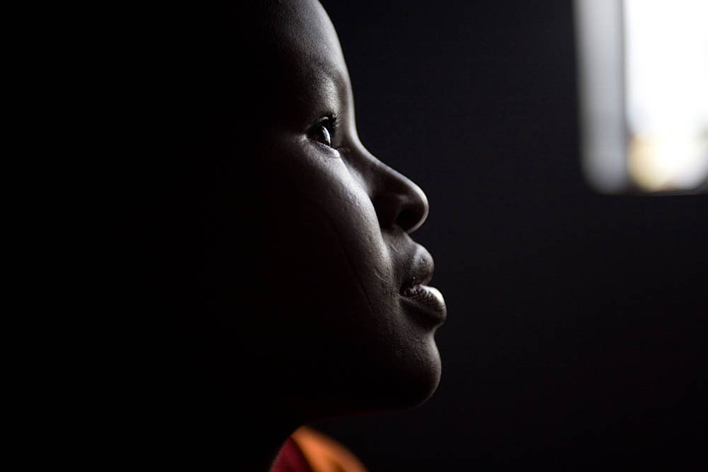 Side profile of a young person with dark skin inside a dark room, illuminated from a window on the right.