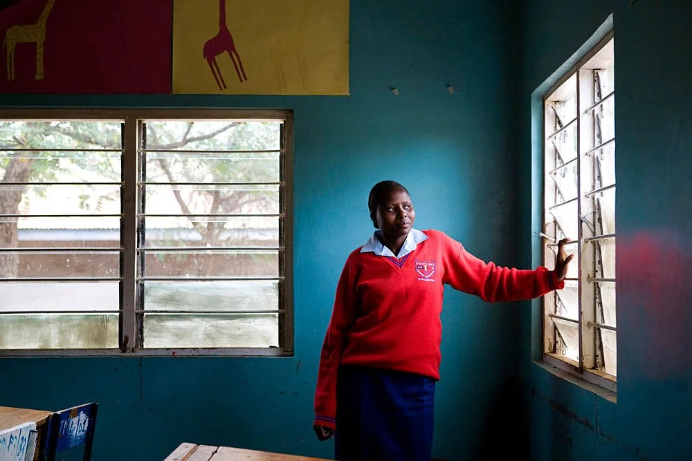 A student in a red school uniform standing inside a classroom, looking out a window with metal bars, while holding the window frame. The classroom has blue walls and a large window, with colorful posters and yellow chairs visible.