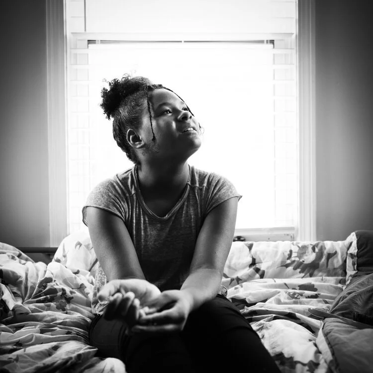 A young woman sitting on a bed, gazing thoughtfully upward, near a window with blinds, in black and white.