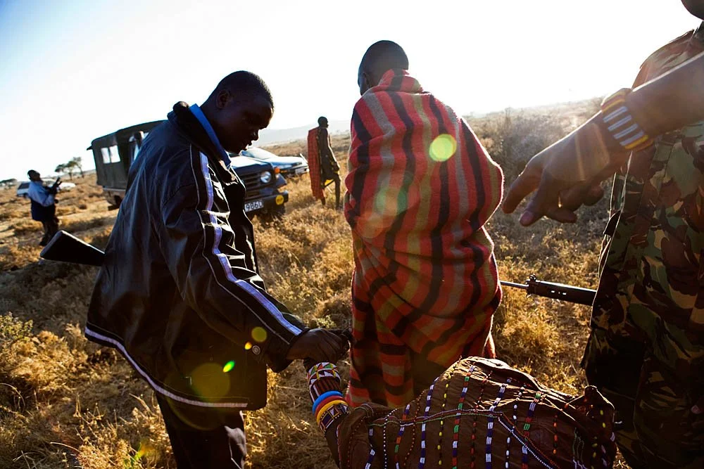 Group of people in an outdoor setting, some dressed in traditional Maasai attire, others in uniforms, engaged in conversation or activity, with vehicles and a dry landscape in the background.