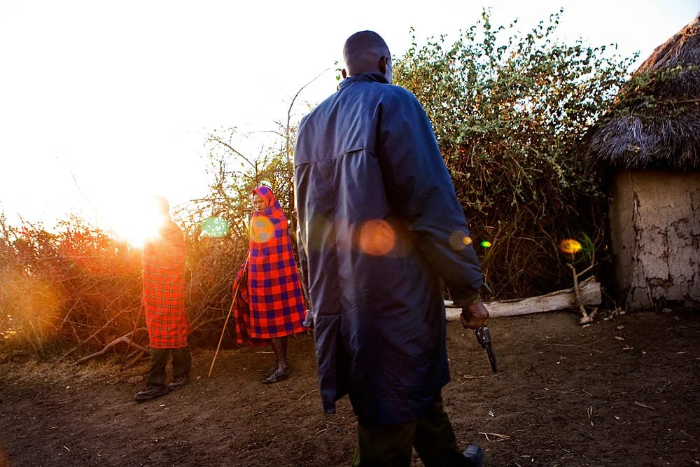 Three Maasai individuals, two women and one man, walking outdoors during sunset; the women are dressed in red and blue shuka cloth and the man is holding a pistol.