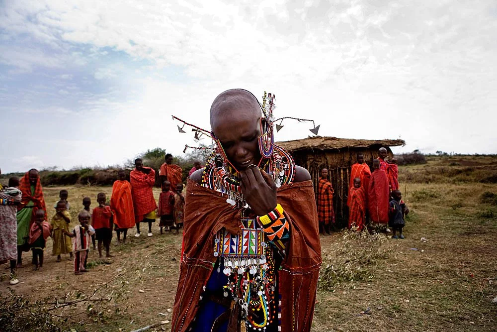 A woman in traditional Maasai attire and jewelry in front of a group of people and a hut in a rural landscape.