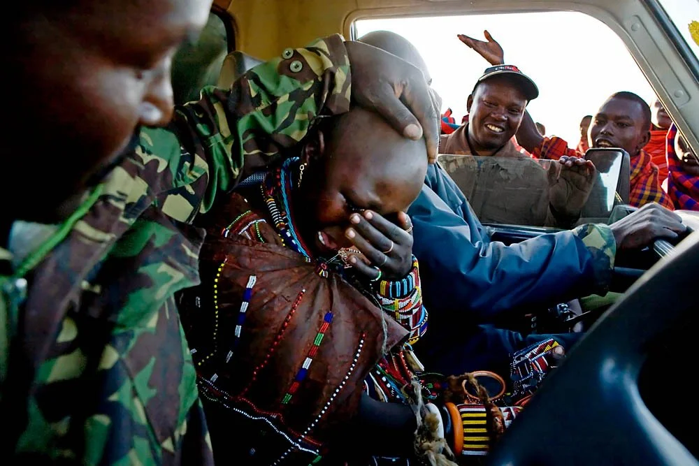 A woman with traditional jewelry inside a vehicle, being comforted by a man with a camouflage jacket, while others look on outside the vehicle, some smiling and taking photos.