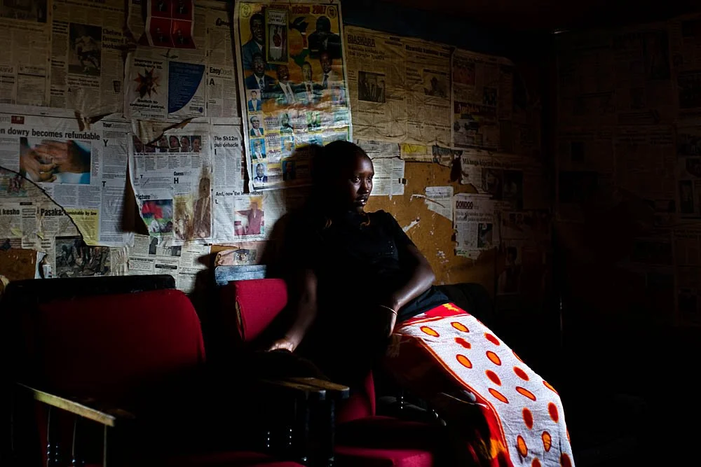 A woman sitting on a red chair, with dark lighting on her face, in a room with walls covered in torn newspapers and posters.