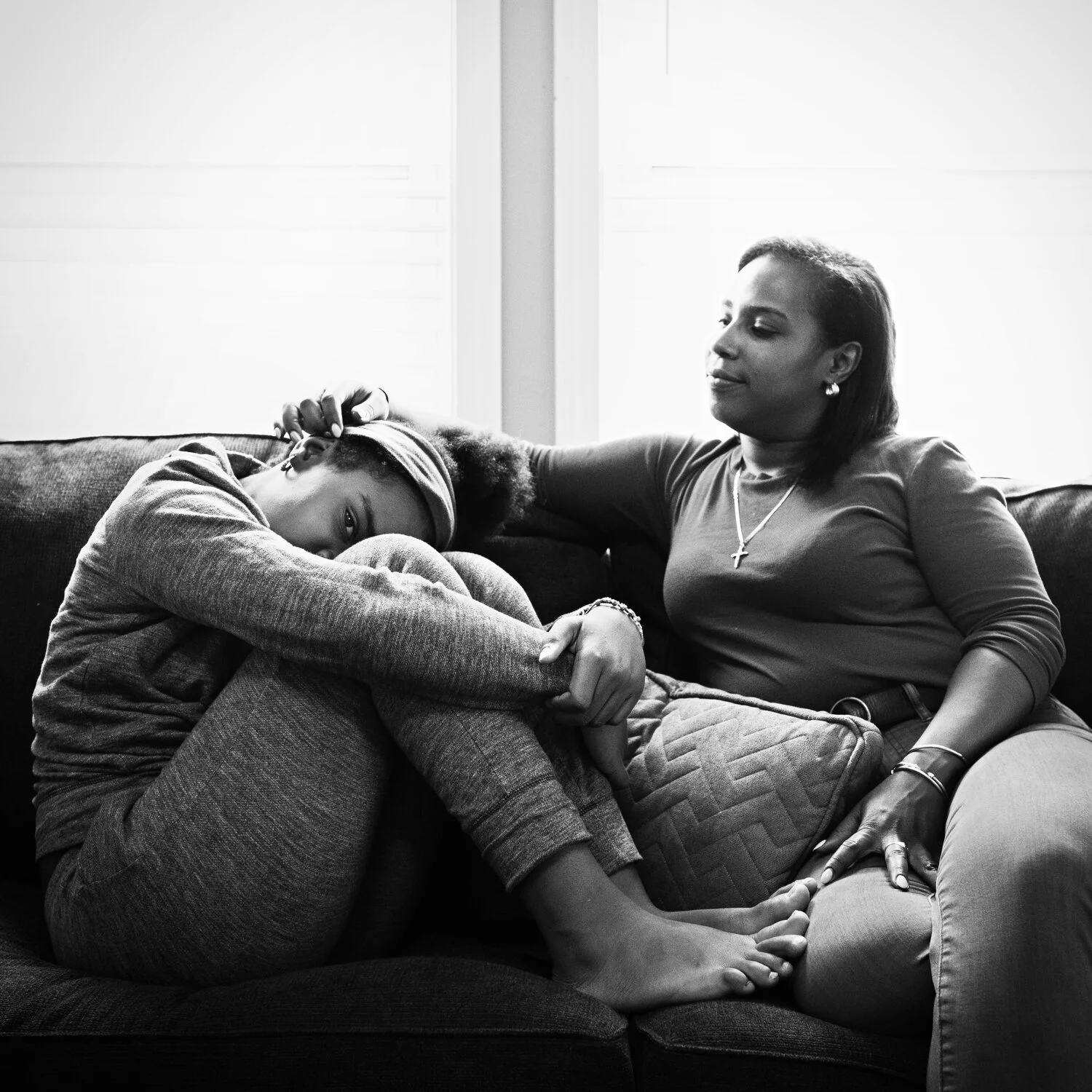 A woman in a short-sleeved shirt and jewelry sitting on a sofa, gently holding a young girl with a headband who is curled up and resting with her eyes open