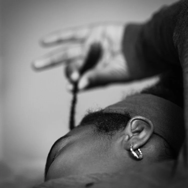 A person with short hair and hoop earrings is resting their head on a surface, while another person is pouring a liquid over their head from a small container.