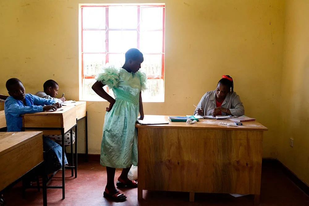 A woman in a light blue dress standing at a desk in a classroom with children seated at desks, while a woman in a white coat sits at her desk with writing materials.