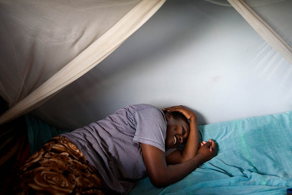 Man lying on a bed, smiling and holding a small object, with his head resting on his hand, inside a tent