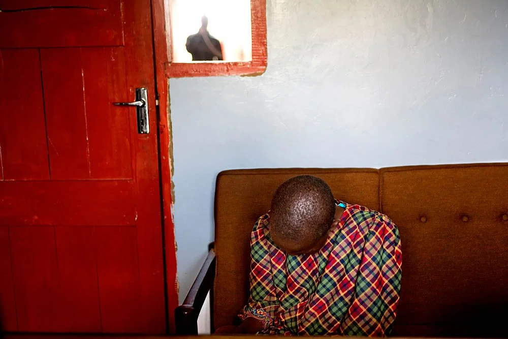 A man with short hair sitting on a brown sofa, wearing a colorful plaid shirt, with his head bowed down. Behind him is a white wall and a closed red wooden door. A person is seen in a small window or opening in the wall, looking into the room.