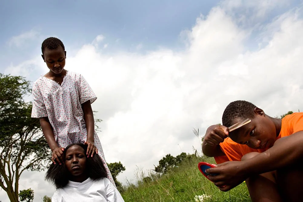 Two children outdoors with one girl standing behind and placing her hands on the head of a girl sitting on the ground, while a boy with a comb and mirror looks at himself.