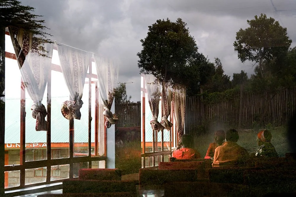 Four people sitting inside a room, looking out large windows with white patterned curtains, overlooking an outdoor area with a wooden fence and trees, on a cloudy day.