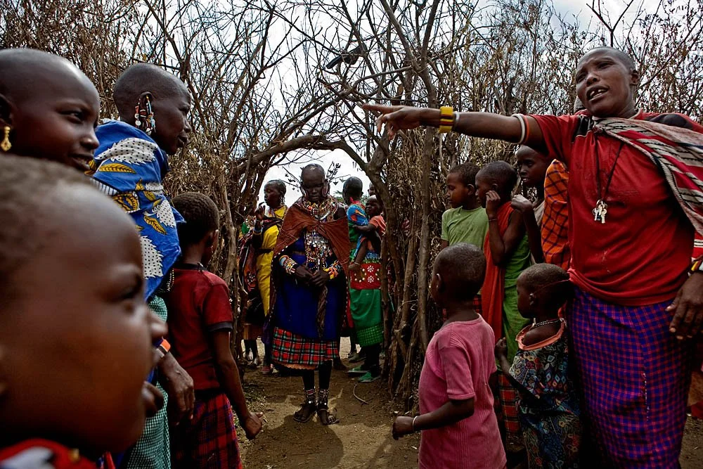 A group of people, mostly children, gathered outdoors among leafless trees, with a woman in traditional attire standing in the center, while others listen to a woman on the right who is gesturing and talking.