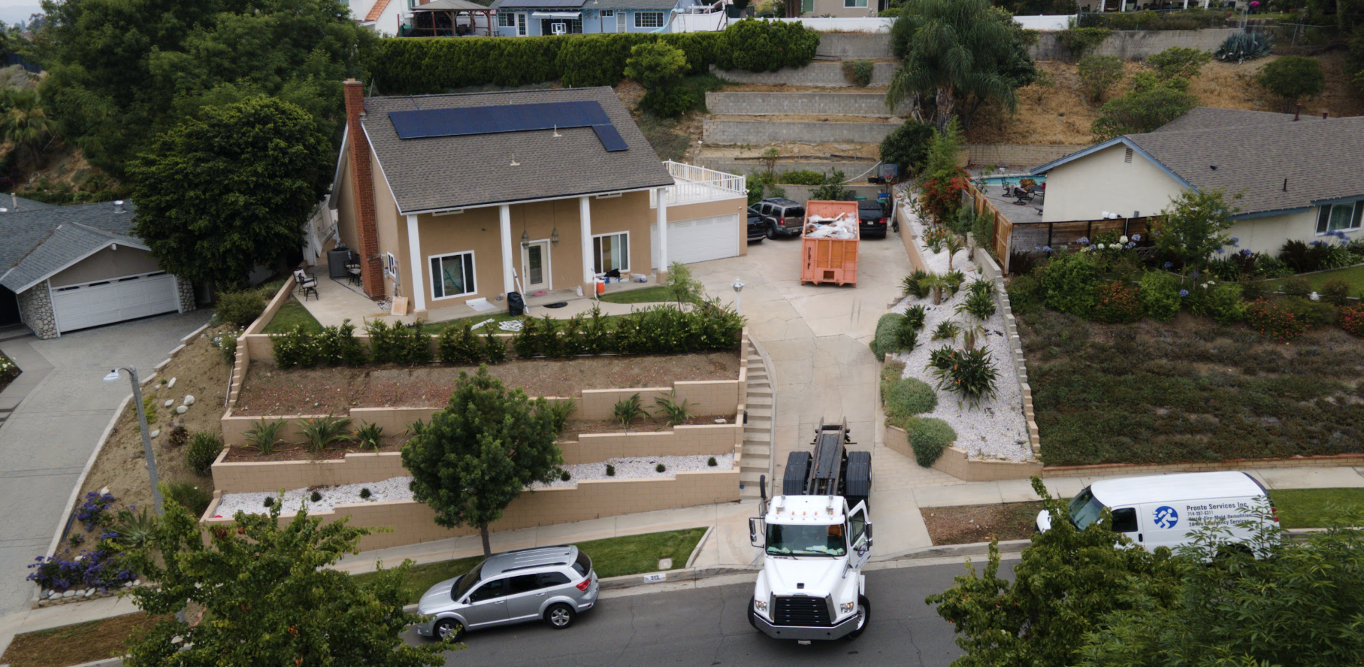 A residential backyard with a house, landscaped yard, construction yard, and vehicles, including a truck, a van, and a car, in a suburban neighborhood.
