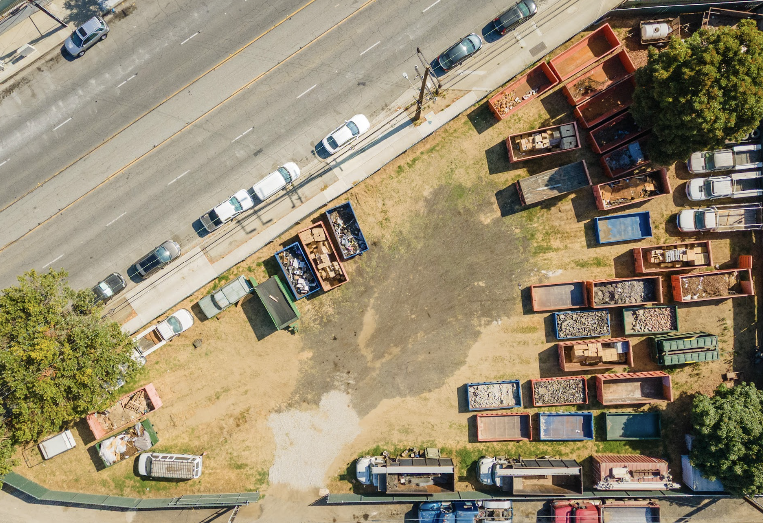 Aerial view of parking lot with parked cars along a city street, next to a fenced area with multiple empty dumpsters filled with construction debris and materials.