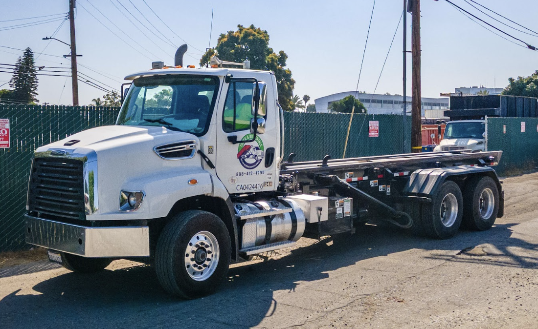 White flatbed truck parked on a street near a construction area, with a fence and utility poles in the background.