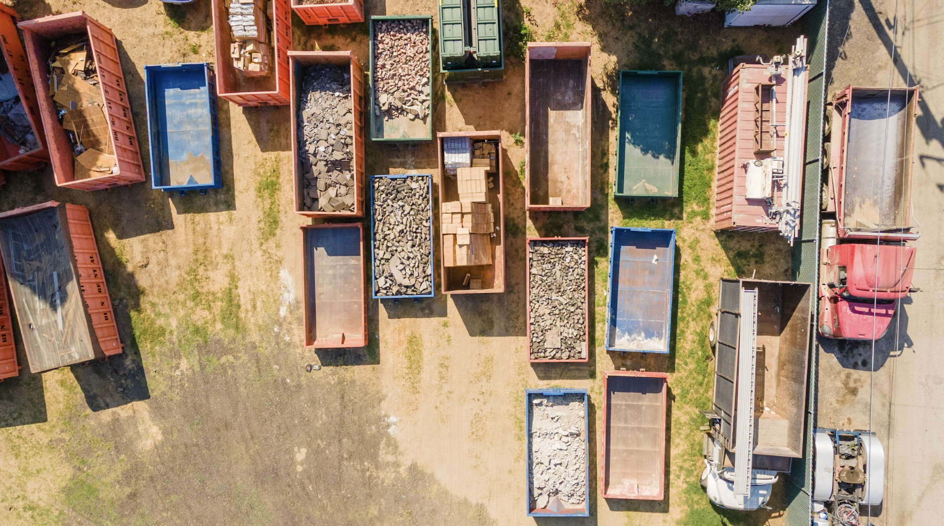 Aerial view of open-top trucks filled with construction materials and debris, parked on a lot with mixed grass and dirt, with vehicles and a fence on the right side.