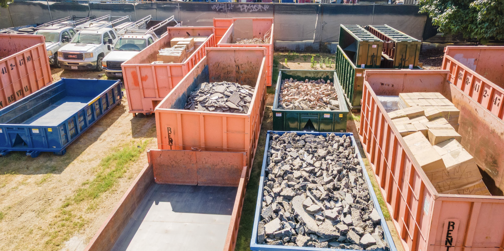 Multiple dumpsters filled with construction debris such as broken concrete, bricks, and tiles, parked in an outdoor lot with trucks and cars in the background.