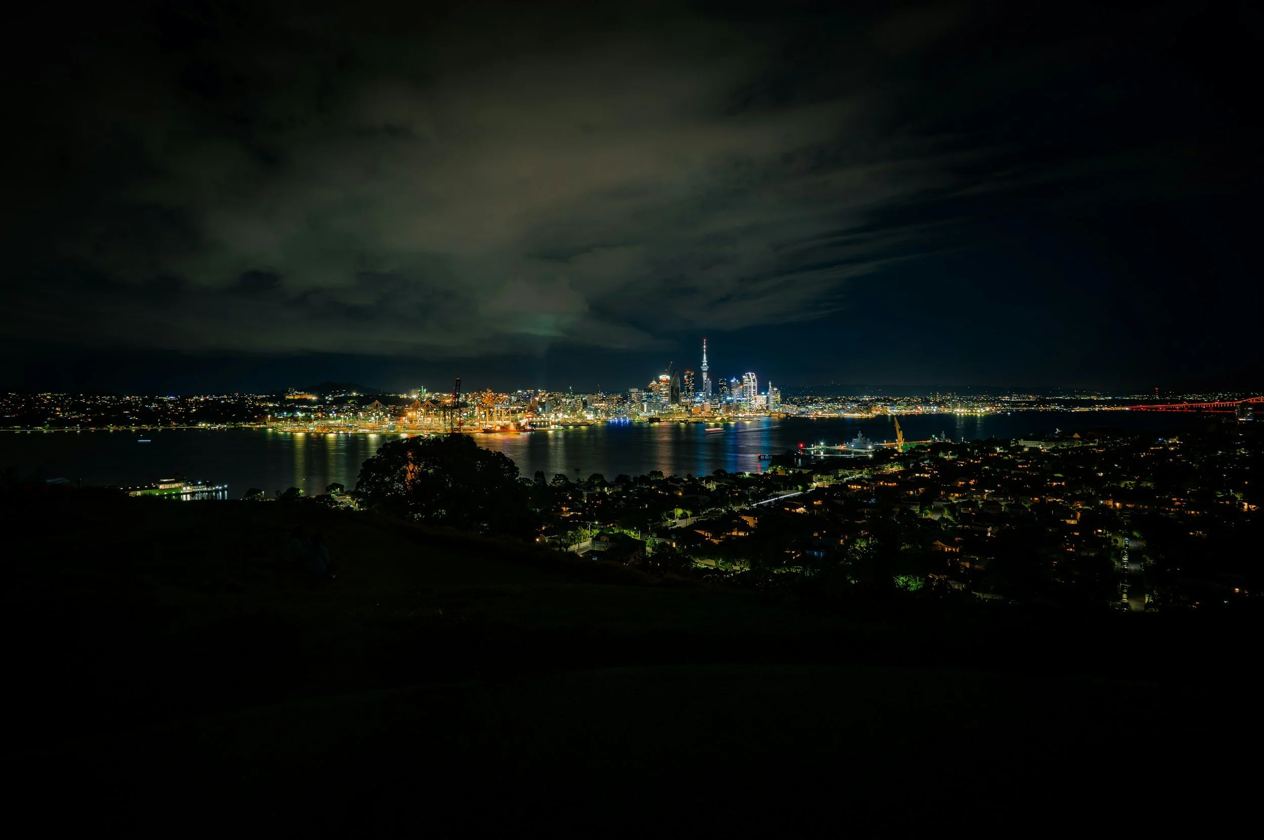 Nighttime view of a city skyline across a body of water, with illuminated buildings, a cloudy sky, and a dark foreground.