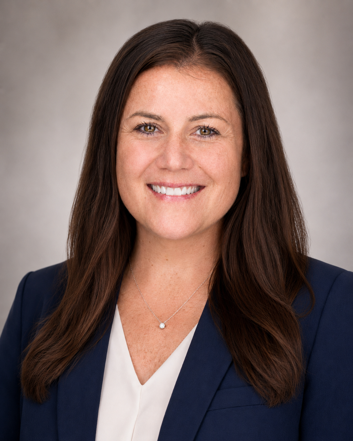 Headshot of a woman with long brown hair, wearing a navy blazer and a white top, smiling and facing forward.
