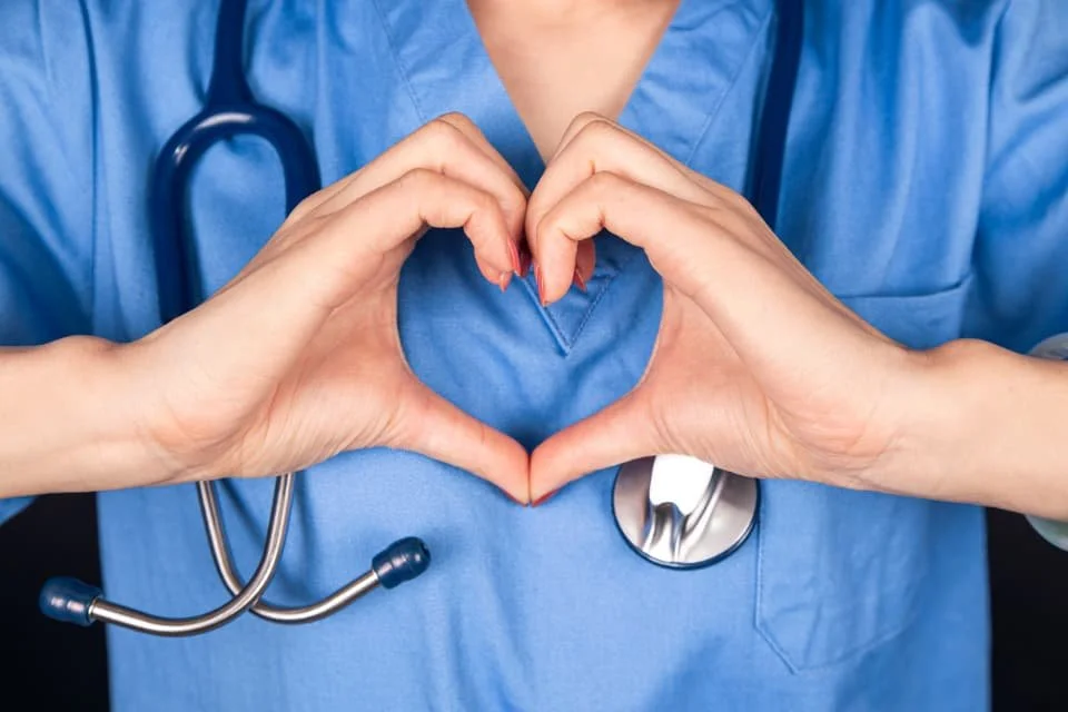 A healthcare professional in blue scrubs forming a heart with their hands, with a stethoscope hanging around their neck.