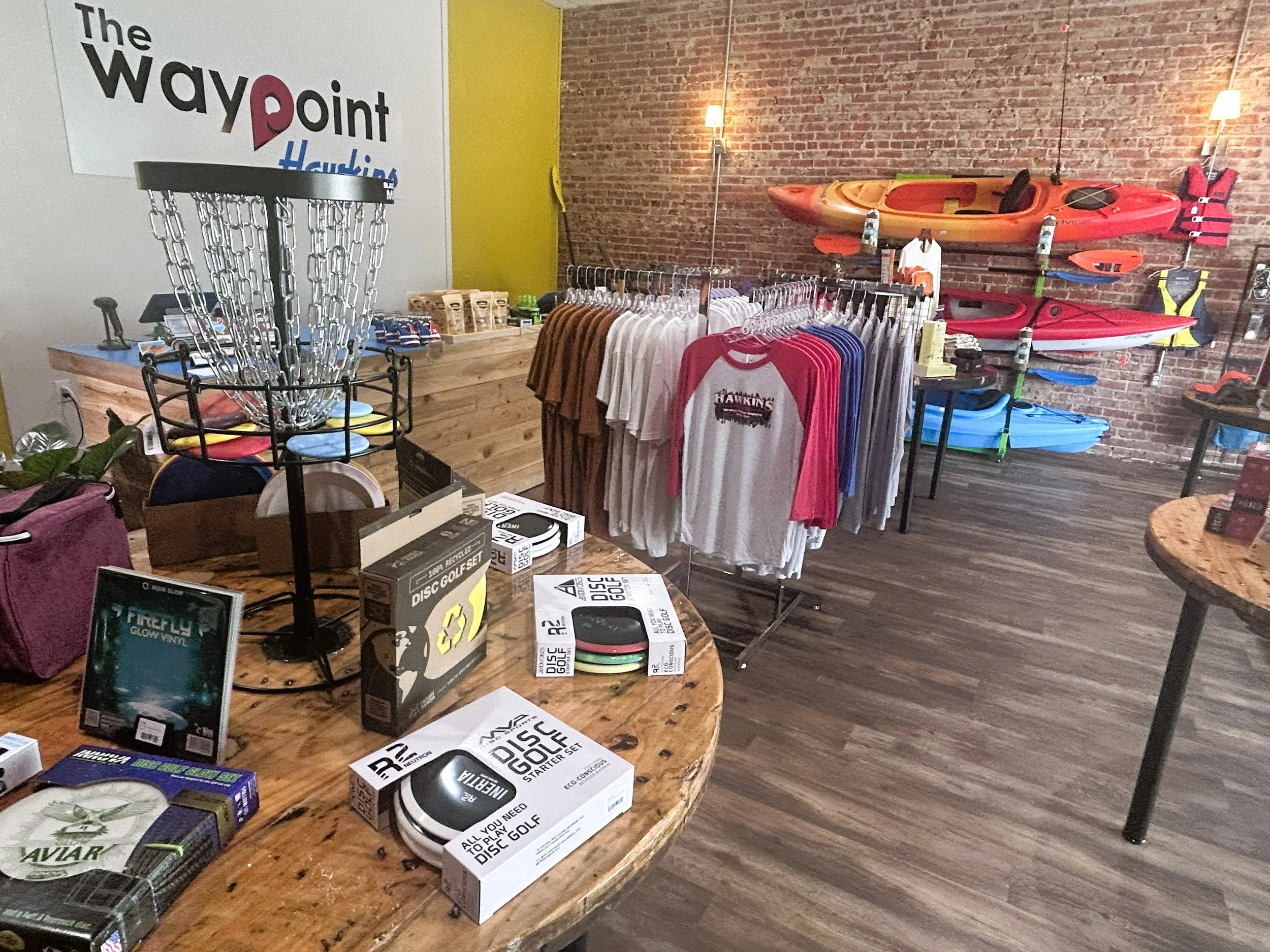 A selection of disc golf starter sets and accessories sit in front of several shirt options with a range of kayaks