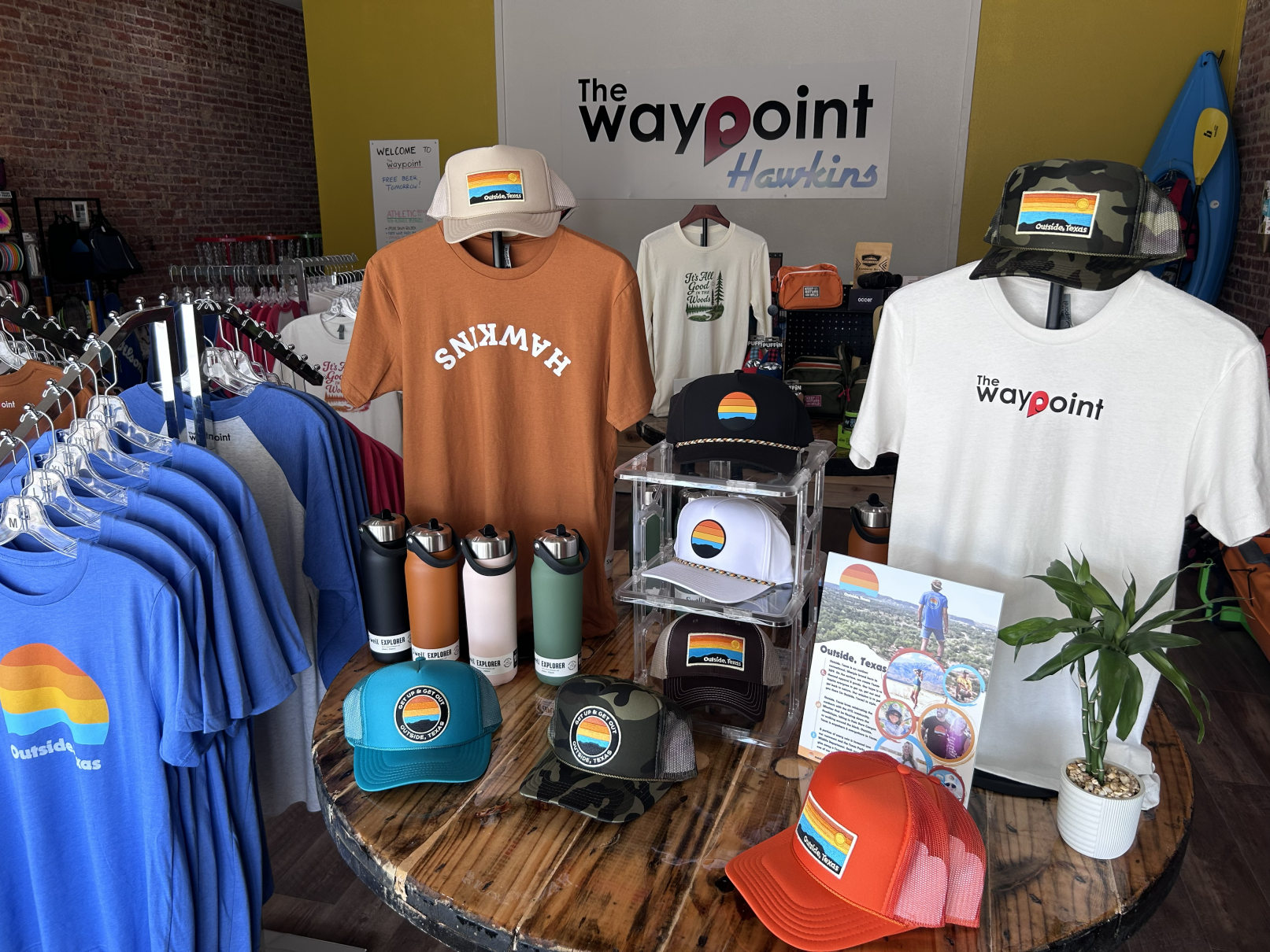 Display of apparel and accessories with outdoor and sunset design, including T-shirts, hats, water bottles, and a potted plant, in a retail store with a sign that reads 'The Waypoint Hawkins'.