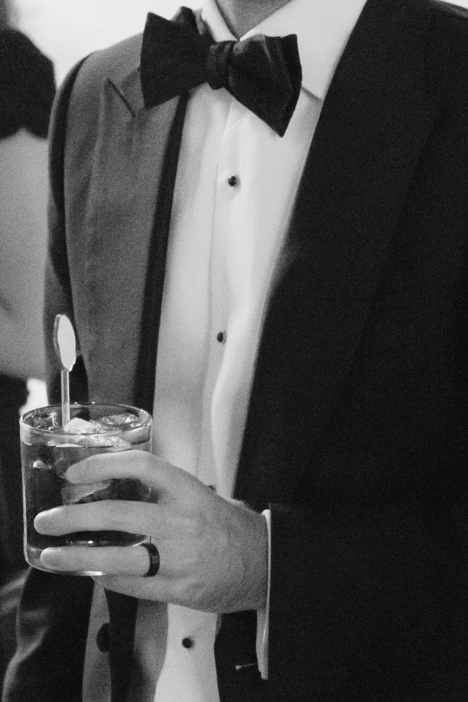 Close-up of a man wearing a tuxedo with a bow tie, holding a glass of cocktail with ice. The image is in black and white.