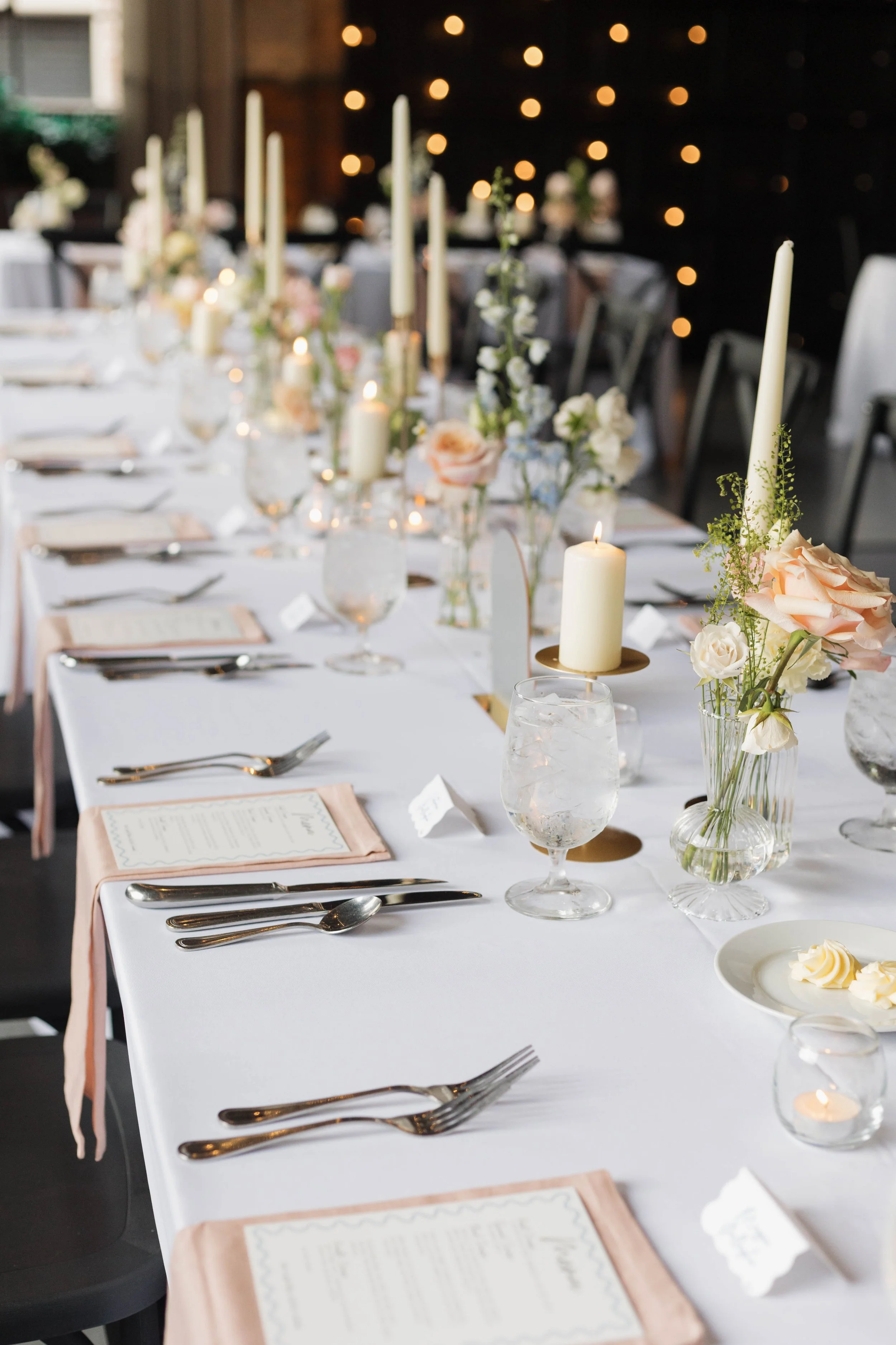 A long wedding reception table decorated with candles, flowers, and place settings with menus and silverware.