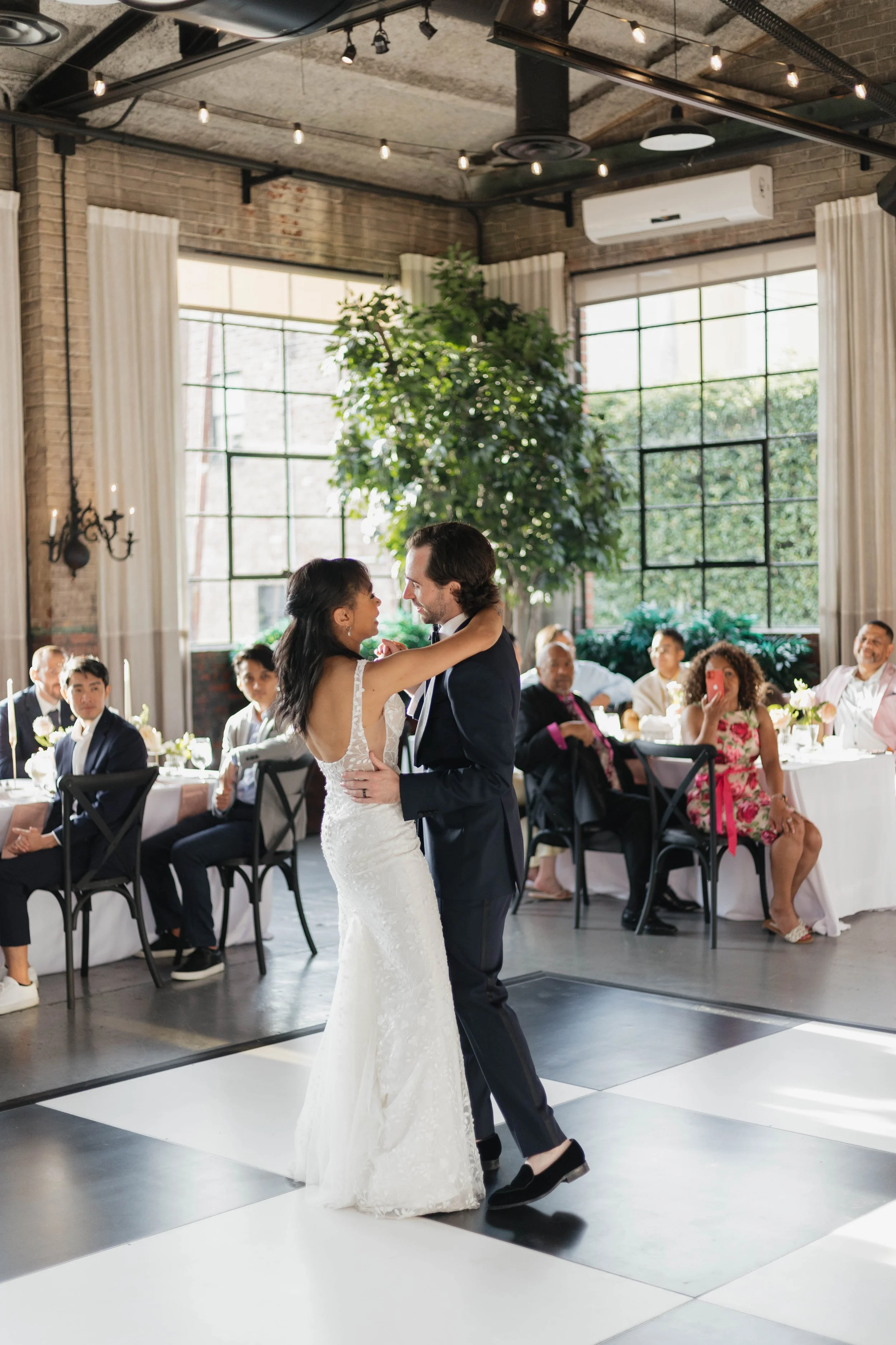 A bride and groom dancing at their wedding reception inside a decorated industrial-style venue with large windows and string lights.