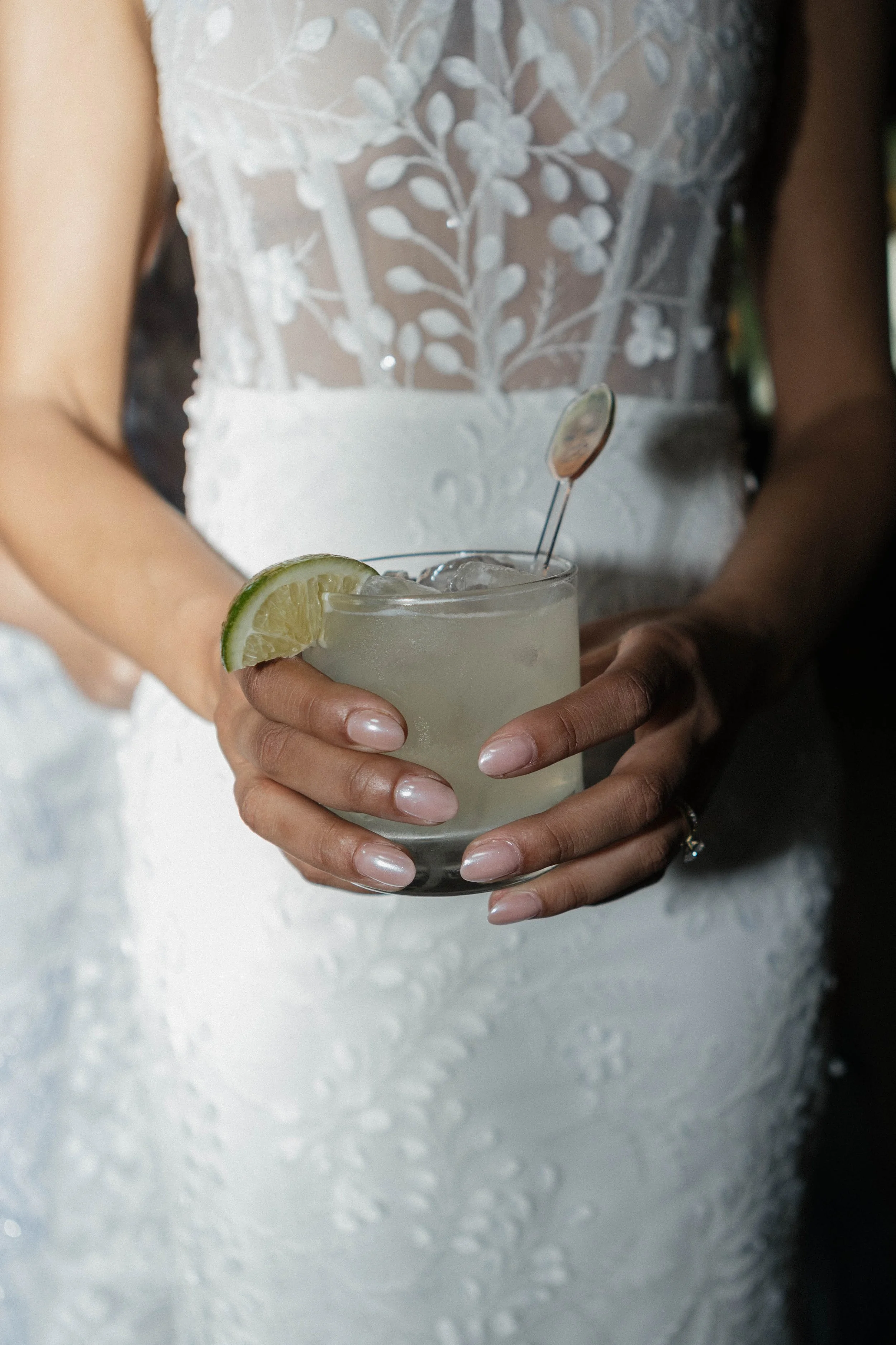 Woman in a white lace wedding dress holding a cocktail glass with lime wedge, ice, and a drink stirrer.