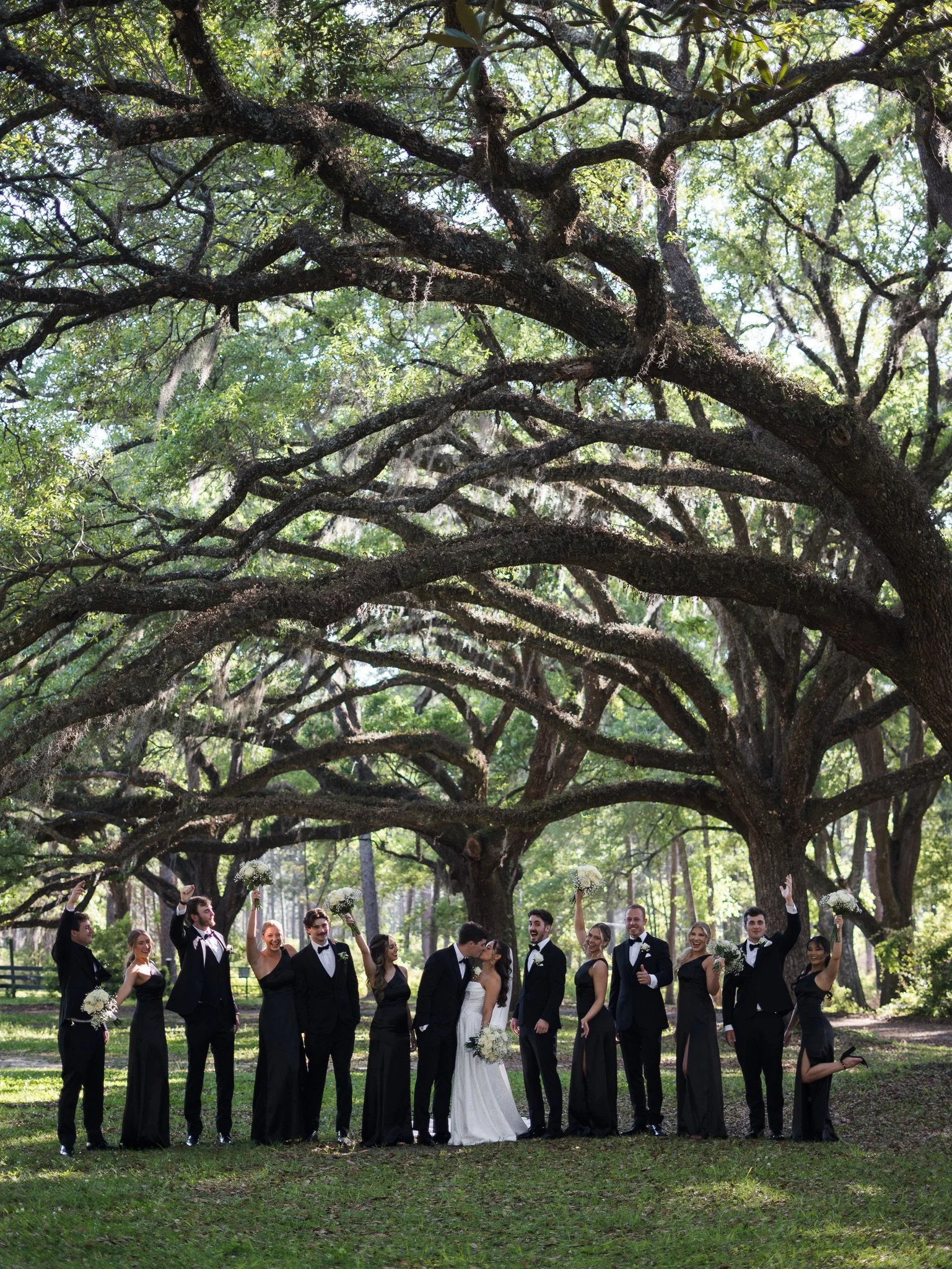 A wedding party of fourteen people standing outdoors under large, sprawling trees with lush green foliage. The bride and groom are kissing in the center, surrounded by bridesmaids and groomsmen dressed in black, some holding bouquets or raising their