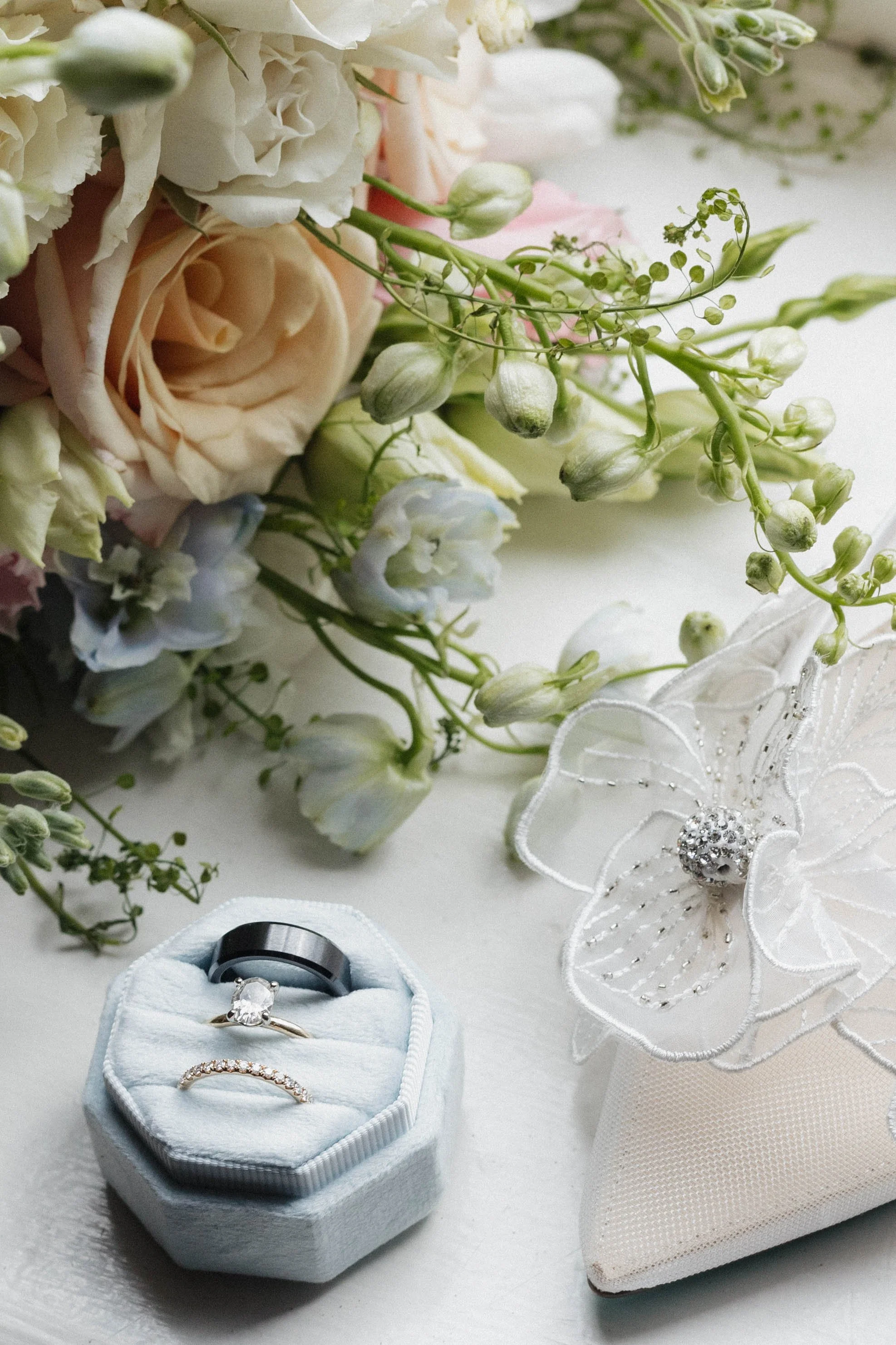 Close-up of wedding rings in a box, a white embroidered flower with bead embellishments, and a bouquet of pastel-colored roses and white flowers.