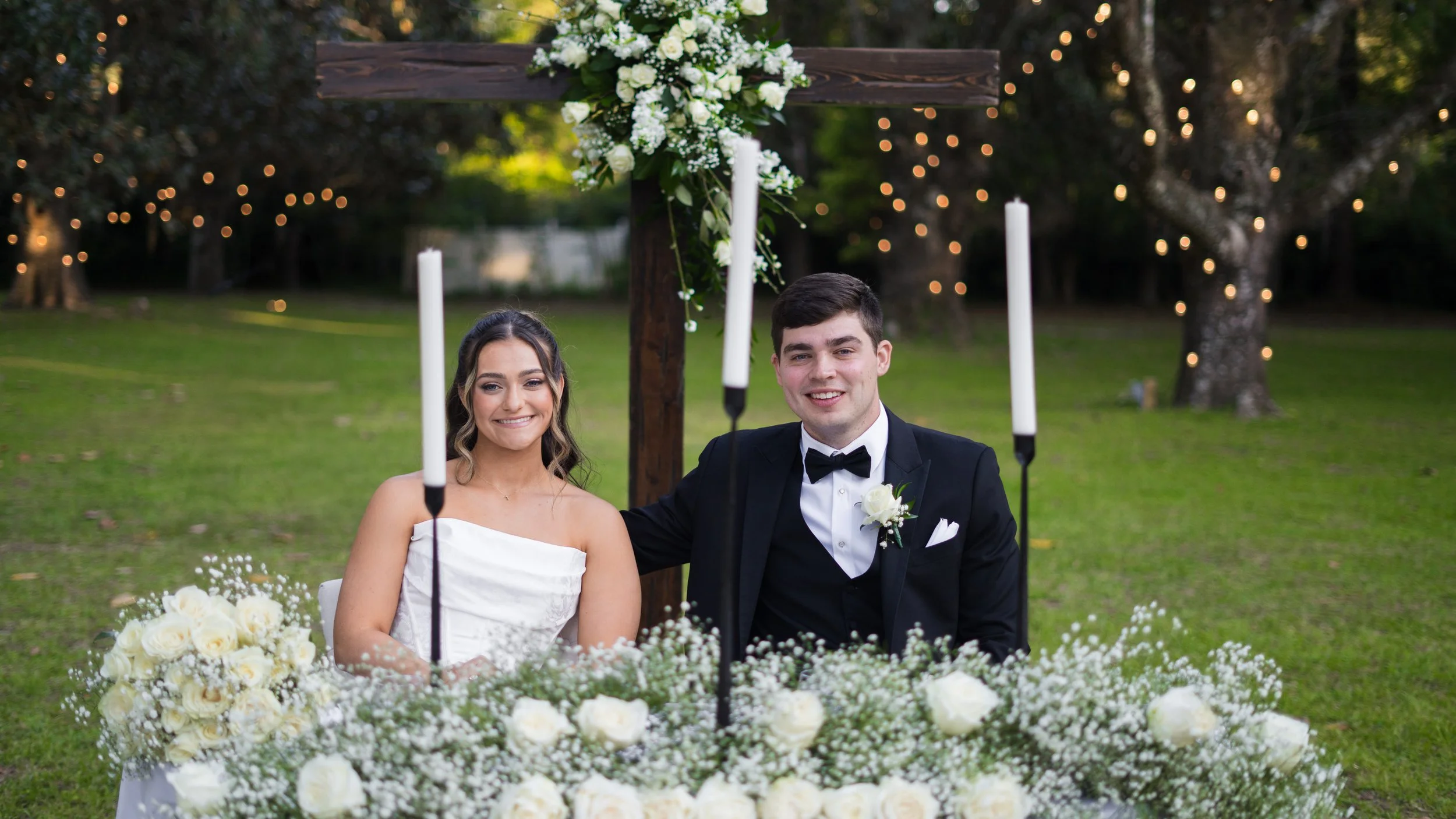 A bride and groom sitting at a decorated outdoor wedding altar with candles, flowers, and trees with string lights in the background.