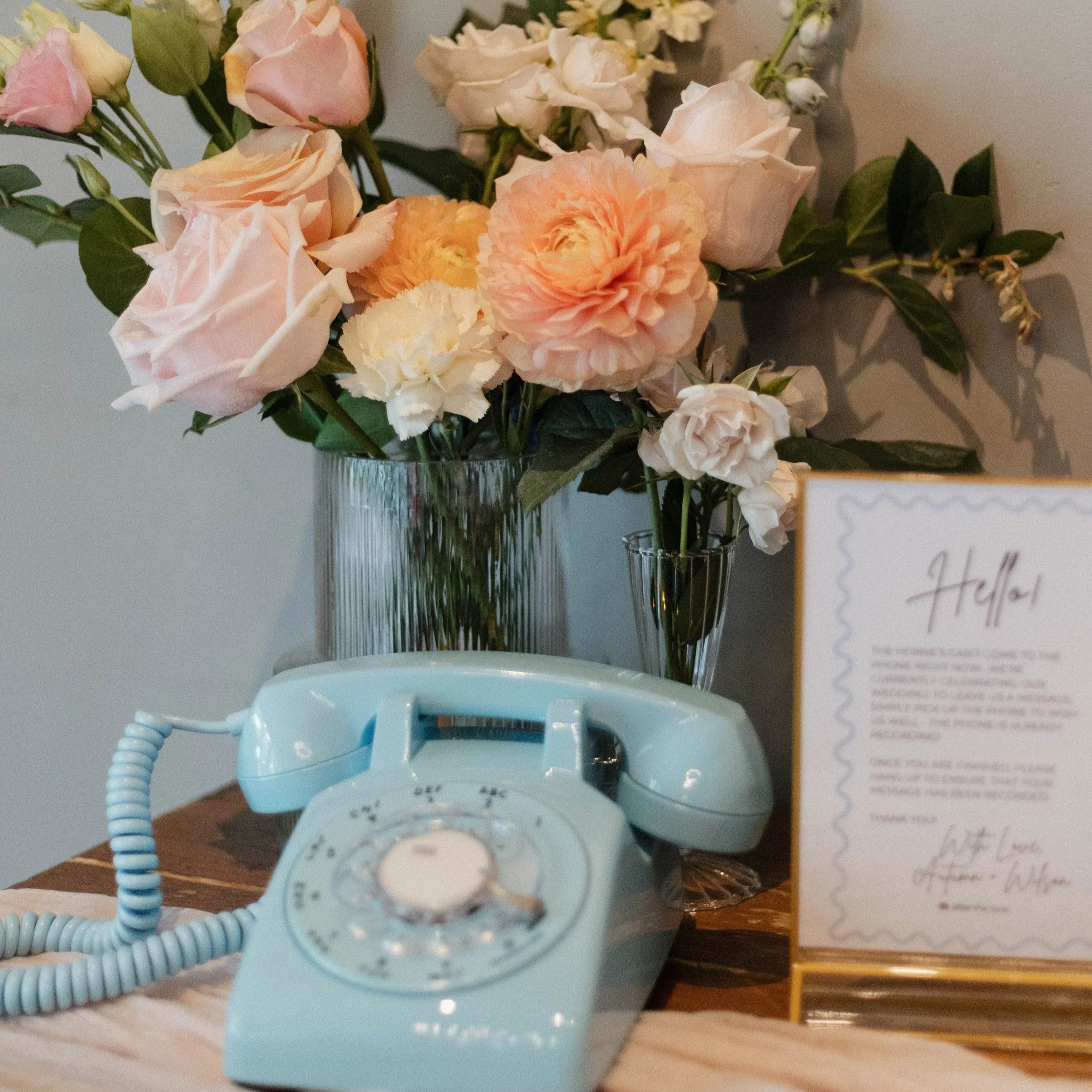 A light blue rotary phone sitting on a wooden surface next to a vase of pink and white flowers and a welcome sign.