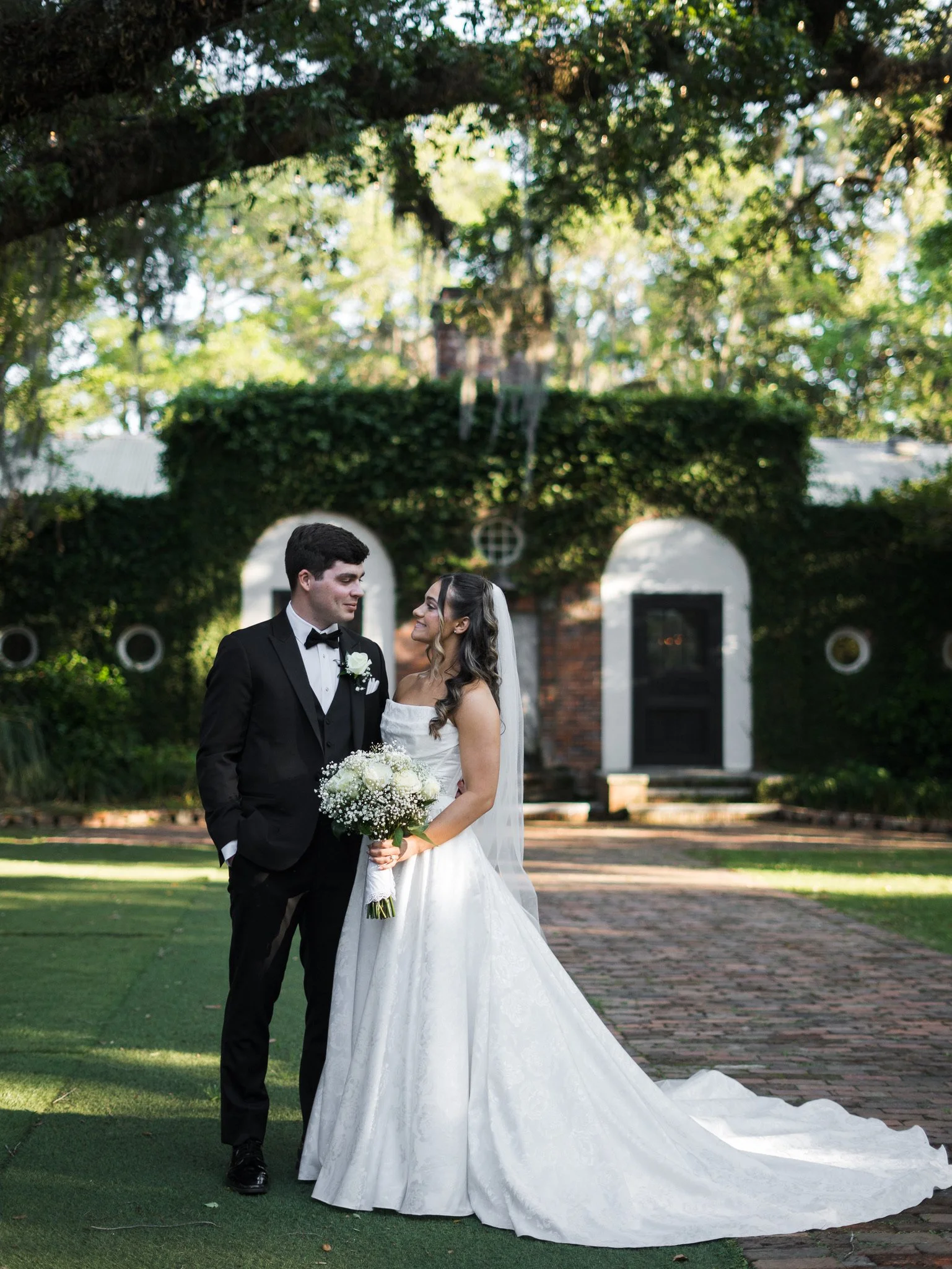A bride and groom standing outdoors on a wedding day, facing each other and smiling. The bride is wearing a white wedding gown and holding a bouquet of white flowers, while the groom is dressed in a black tuxedo with a bow tie.