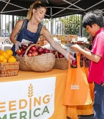 A woman at a market stall handing a bag of produce to a young boy, with a sign that reads 'Feeding America' and baskets of fruit on the table.