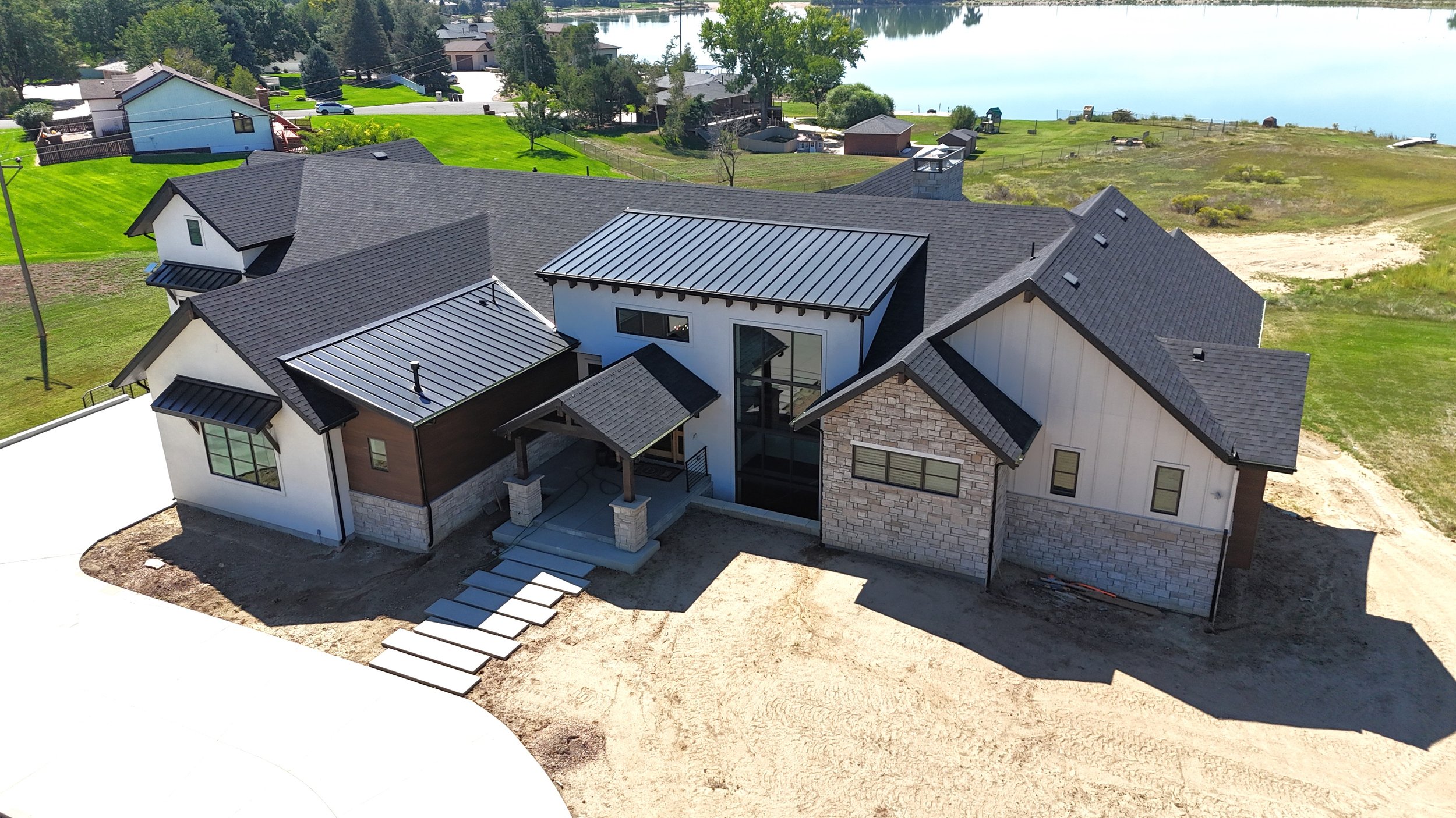 Aerial view of a modern house with black metal roofs, white and brown exterior walls, and large windows, situated near a body of water with surrounding green lawns and neighboring houses.