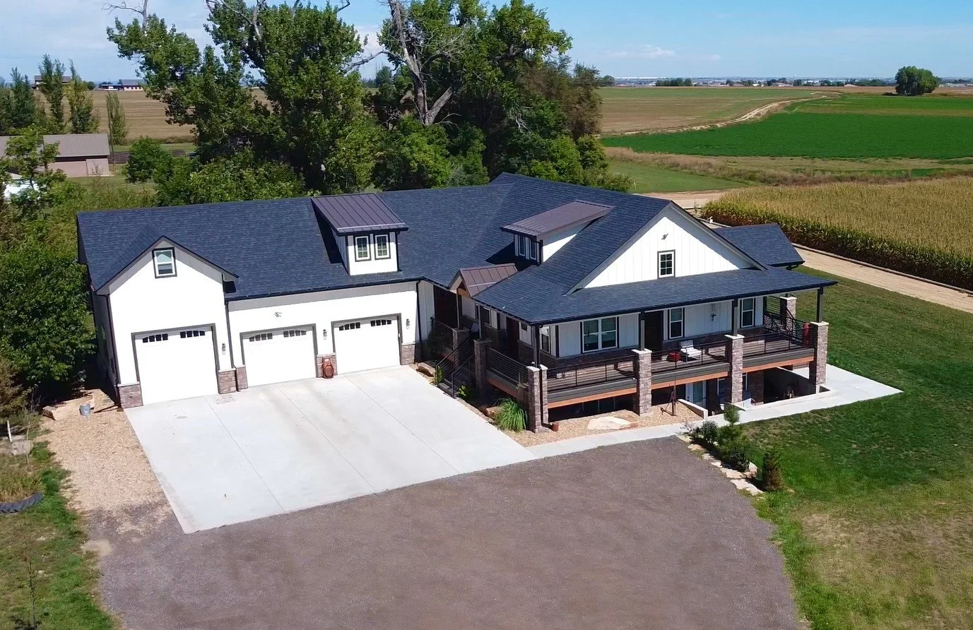A large, modern house with a black roof, white walls, and a spacious front porch, situated in a rural area with expansive farmland and fields in the background.