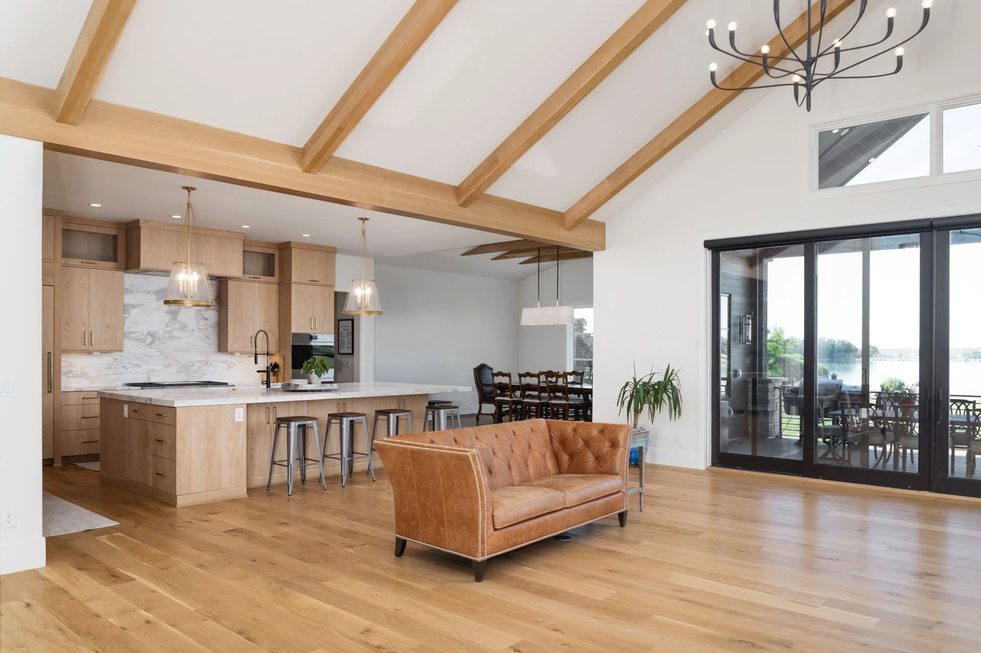 Open-concept living space with a kitchen, dining area, and a sitting area. The kitchen has light wood cabinetry, a marble backsplash, and pendant lighting. There is a brown leather tufted sofa, hardwood floors, and large sliding glass doors leading t