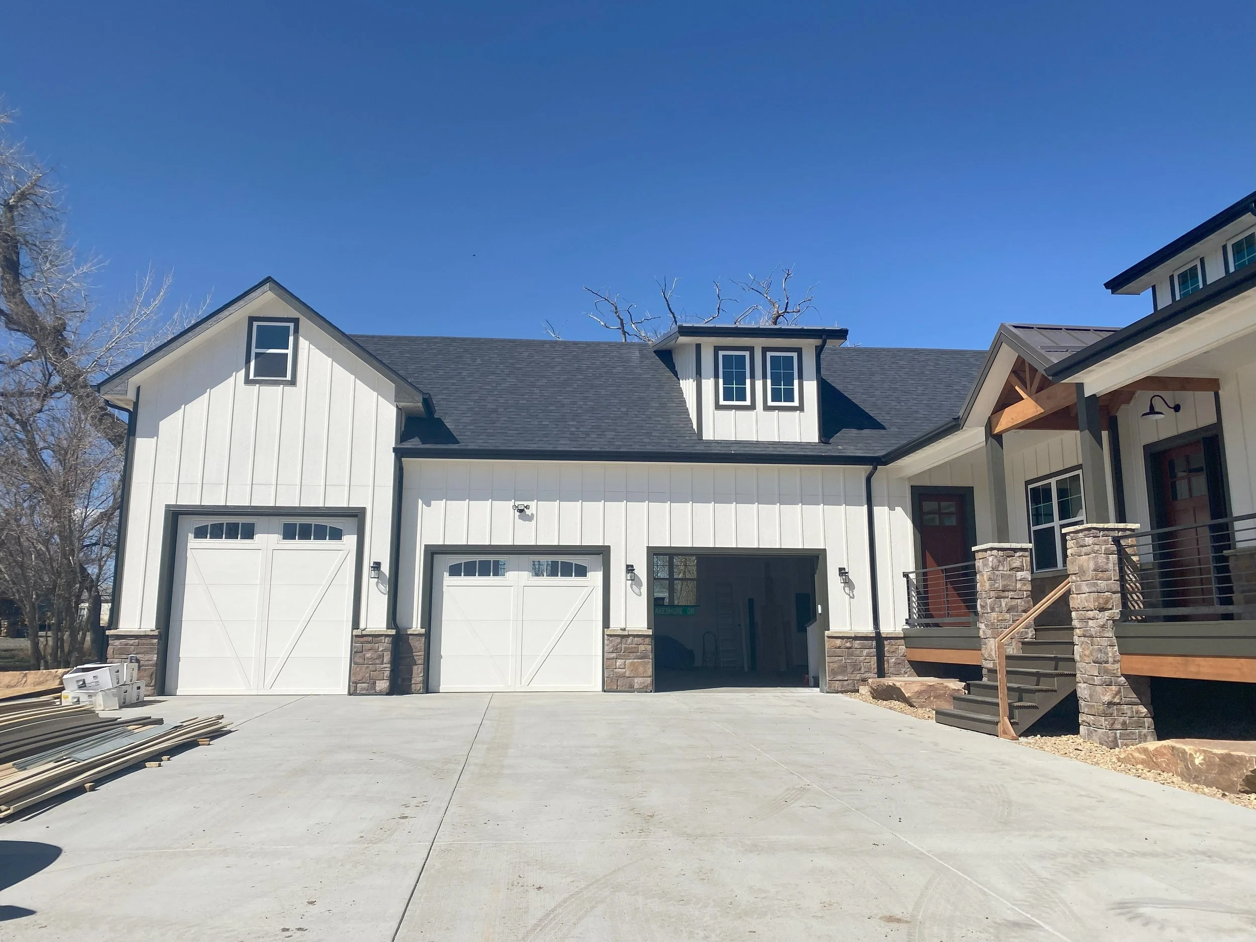 Newly constructed modern house with white siding, stone accents, black roof, and two garage doors on a clear day.