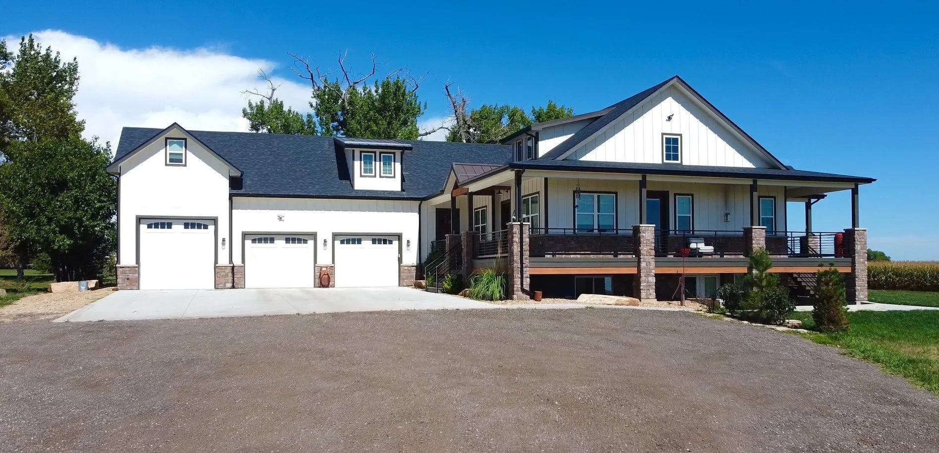 A modern house with a large front porch, white exterior, black roof, and multiple windows. There is a three-car garage attached on the left side, with a gravel driveway in front. The house is surrounded by trees and a partly cloudy blue sky.
